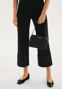 Črna prešita torbica z zlatim zadrgo in logotipom, kombinirana s črnimi skrajšanimi hlačami ter črnimi balerinkami z sijočim prstnim delom in pentljasto okrasjo.