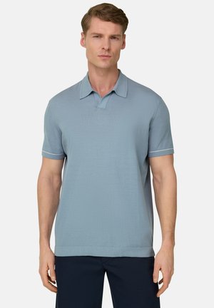 Homme portant un polo à manches courtes bleu clair avec bordure blanche sur les manches et un pantalon foncé, debout devant un fond uni.