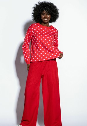 Czerwony sweter z różowym wzorem serc, w połączeniu z szerokimi czerwonymi spodniami o prążkowanej fakturze. Materiał wydaje się miękki i teksturowany.