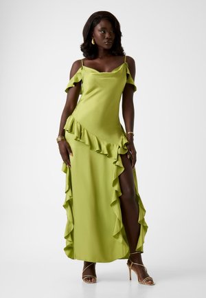 Mujer de piel oscura y cabello ondulado, vestida con un vestido verde lima con volantes, sin hombros y con una abertura alta en el muslo, y tacones dorados con tiras.