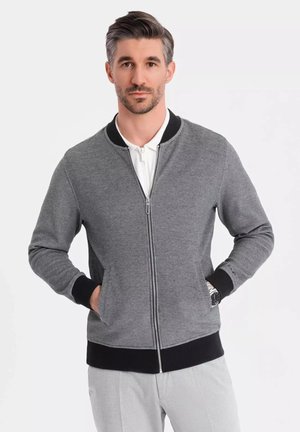 Chaqueta gris con cremallera y acentos negros, con puños y cinturilla acanalados. Presenta bolsillos laterales y una textura suave.