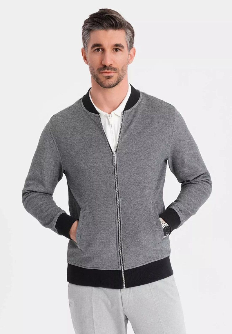 Šedá zip-up bunda s černými akcenty, žebrovanými manžetami a páskem. Má boční kapsy a hladkou texturu.