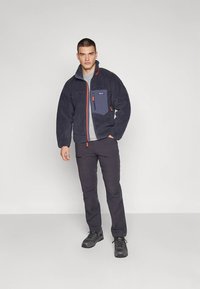 Marinblå fleecetröja med en ljusgrå panel och bröstficka, orange dragkedjedetalj, kombinerad med mörka byxor och svarta vandringsskor.