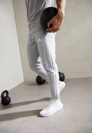 Personne portant un pantalon de survêtement Adidas gris et des baskets blanches tenant un ballon médicinal noir dans une salle de sport avec des kettlebells au sol.