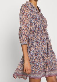 Blommig klänning i marinblått med persika- och vita blommor, midjemarkering, trekvartsärmar och mönstrad fåll. Tillverkad av lättviktig tyg.
