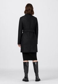 Mustapukuinen, räätälöity takki, jossa on vyö, pitkät hihat ja jäsennelty siluetti, yhdistetty polven yläpuolelle ulottuviin kiiltäviin mustiin saappaisiin.