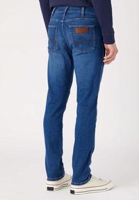 Jeans de mezclilla, de corte ajustado, azules oscuros, con un detalle de parche de cuero en la parte trasera, costuras en contraste y una textura suave. Llevados con zapatillas blancas.