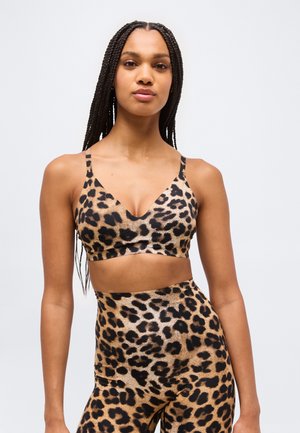 Ženska z dolgimi spetimi lasmi, oblečena v športni modrček s potiskom leopardove kože in ujemajoče legice, stoji pred enostavnim svetlim ozadjem.