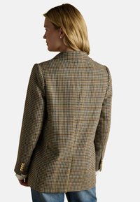 Sako s houndstooth vzorom v hnedej, krémovej a bordovej farbe s priliehavým strihom, klopovým golierom a zlatými gombíkmi na rukávoch.