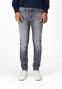 Muž v zesvětlených šedých skinny džínách a bílých teniskách, stojící na bílém pozadí, s rukama u těla.