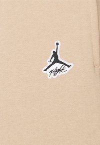 Beige sweatshirtstof med et sort Air Jordan-logo-patch med hvid kant, der viser en springende figur og ordet "Flight."