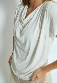 Vit, draperad t-shirt med korta ärmar, mjukt tyg och cowl-halsringning. Bärs tillsammans med neutrala byxor som har en subtil textur och avslappnad passform.
