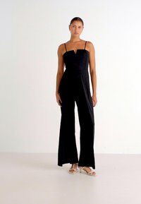 Macacão preto com alças finas, design em V e calças largas. Usado com sandálias de salto branco de ponta aberta. Textura de tecido suave.