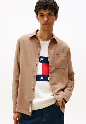 Brown button-up shirt made of soft fabric, featuring a chest pocket and paired with a cream sweater displaying a logo in red, blue, and white.
