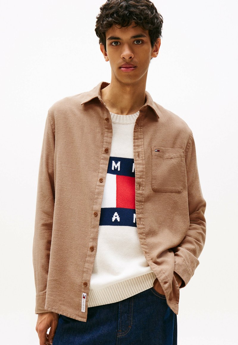Brown button-up shirt made of soft fabric, featuring a chest pocket and paired with a cream sweater displaying a logo in red, blue, and white.