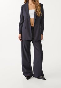 Laivastonsininen raidallinen puku, jossa on muotonsa mukainen pitkä bleiseri, jossa on kaksi taskua, sekä leveälahkeiset housut. Materiaali näyttää sileältä ja pehmeältä.