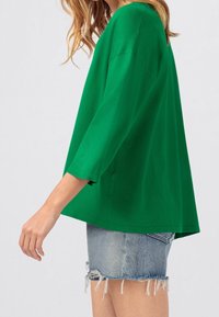 Vrouw in een loszittend felgroen shirt met driekwart mouwen, gecombineerd met een versleten denim minirok.