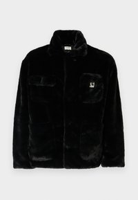 Chaqueta de pelaje sintético negro con un collar, dos bolsillos frontales y un pequeño parche con logo en la parte superior izquierda. Textura suave y ajuste relajado.