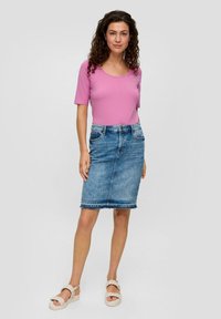 Vrouw met krullend haar in een roze top met korte mouwen, een spijkerrok tot de knie en witte gevlochten sandalen, staand tegen een neutrale achtergrond.