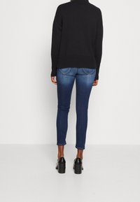 Personne portant un jean skinny bleu délavé, des chaussures à talons carrés noires et un pull noir à manches longues, debout de dos sur un fond uni.