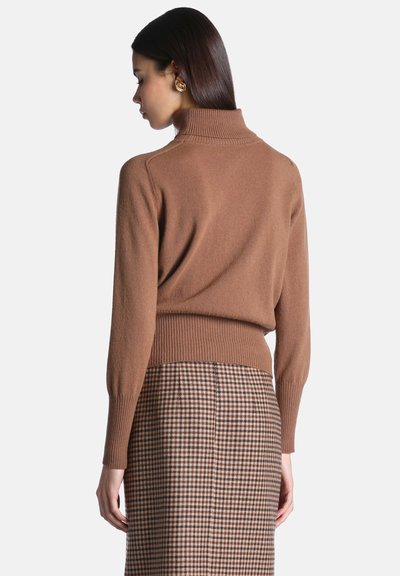 Femme portant un pull à col roulé marron et une jupe marron et noire à carreaux, vue de dos sur un fond uni.