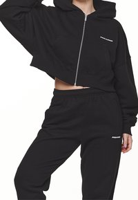 Musta lyhyt huppari, jossa on vetoketju edessä, suuri etutasku ja joustava helma; yhdistetty samansävyisiin mustiin collegehousuihin, joissa on logoyksityiskohta.