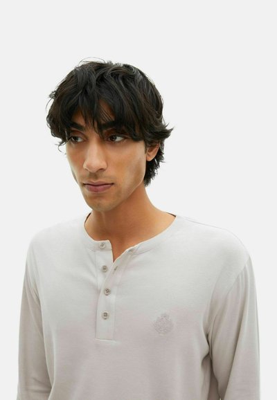 Junger Mann mit schulterlangem, dunklem Haar, der ein hellbeiges Henley-Shirt mit vier Knöpfen und einem dezenten, gestickten Wappen auf der Brust trägt.
