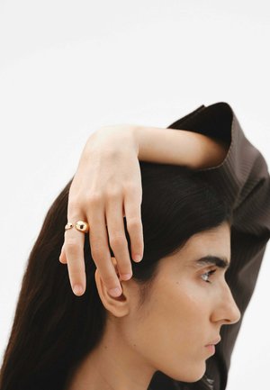 Anillo de oro con formas redondeadas, uno grande y otro más pequeño, llevado en los dedos. Tono de piel visible; mano posicionada sobre la cabeza, mostrando uñas naturales.