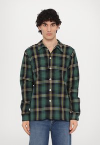 Grønn og svart rutete skjorte med knapper laget av flanell, med gule detaljer, krage og lange ermer. Modellen har på seg jeans.