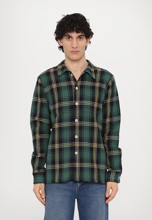 Groen en zwart geruite button-up shirt van flanel, met gele accenten, een kraag en lange mouwen. Model draagt jeans.