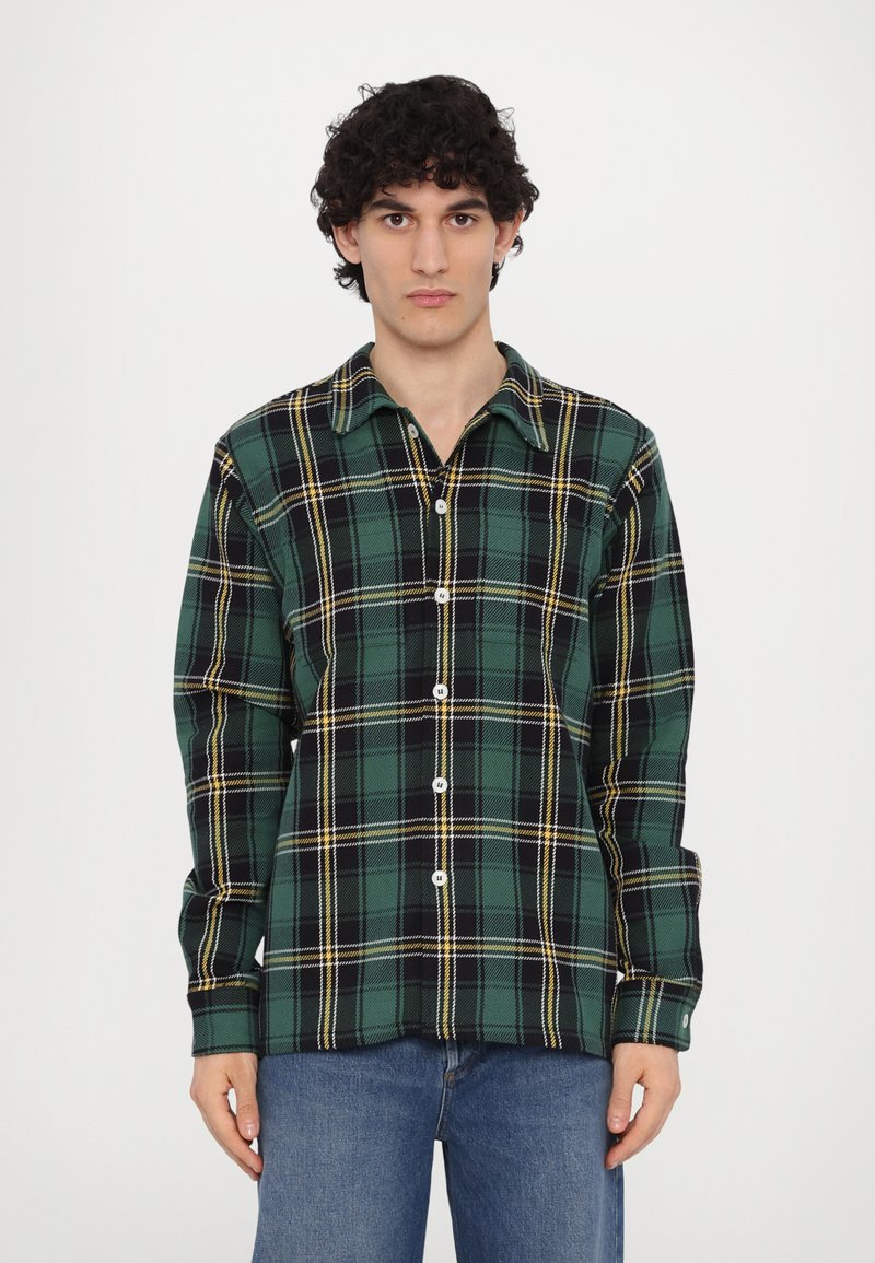 Grønn og svart rutete skjorte med knapper laget av flanell, med gule detaljer, krage og lange ermer. Modellen har på seg jeans.