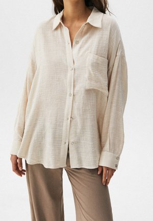 Kobieta w luźnej, kremowej koszuli na guziki z kieszenią na piersi i długimi rękawami, w parze z taupe spodniami na jednolitym tle.