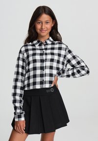 Giovane donna che indossa una camicia a quadri bianca e nera con bottoni e una gonna plissettata nera, che posa con una mano sulla vita.