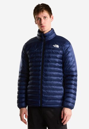 Ung mand iført en marineblå quiltet North Face-jakke med høj krave, stående mod en ensfarvet lys baggrund.