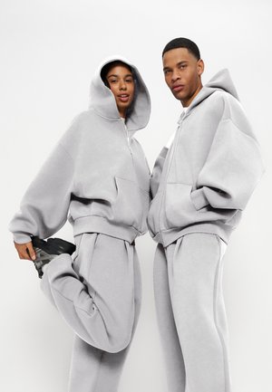 Dos personas con sudaderas con capucha gris claro de gran tamaño y pantalones deportivos a juego, de pie frente a un fondo blanco liso.