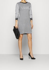 Graues Strickkleid mit schwarzen Akzenten, das ein Logo auf der Brust trägt, lange Ärmel und eine enganliegende Form hat. Kombiniert mit einer schwarzen Clutch und hohen Schuhen.