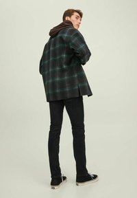 Overdimensjonert svart og grønn rutete jakke med hette, båret over mørke jeans og svarte joggesko. Stoffet fremstår som mykt med en strukturert finish.