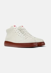 Sneaker alte in pelle bianca con suole in gomma marrone. Presentano lacci rotondi e etichette rosse sulla linguetta. Texture liscia e cuciture sottili.