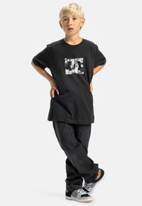 Camiseta de algodón negra con un logo DC blanco desgastado. Combinada con pantalones negros holgados y zapatillas grises con acentos naranjas.