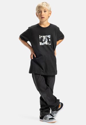 Zwarte katoenen T-shirt met een verweerde witte DC-logo. Gecombineerd met losse zwarte broek en grijze sneakers met oranje accenten.