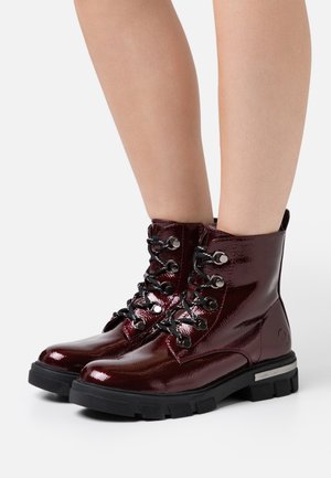 Bottines en cuir verni bordeaux avec des lacets noirs, à bout rond, semelle en caoutchouc et accents métalliques. Hauteur atteignant le mollet.