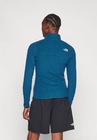 Chaqueta de forro polar azul con cuello alto, mangas largas y un logo en la parte posterior, combinada con pantalones cortos negros con bolsillos laterales.