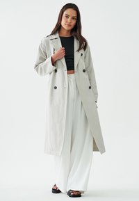 Heledhalli trench coat topelt nööpidega, laiad varrukad ja lahtine lõige, kantud musta lühikese särgi ja valgete laia säärega pükste üle.