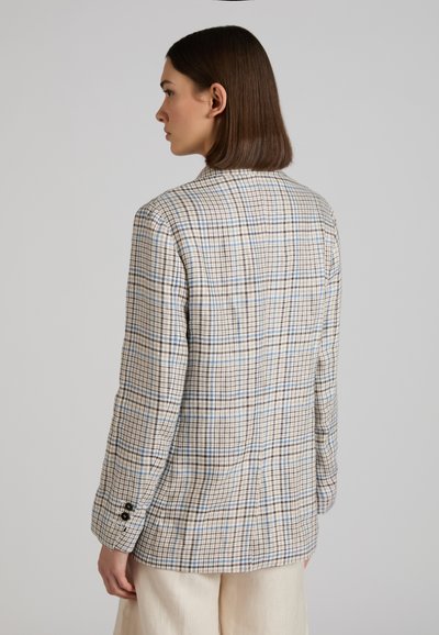 Vrouw van achteren gezien, gekleed in een beige geruit colbert met blauwe en bruine lijnen en loszittende, lichtgekleurde broek, tegen een effen achtergrond.