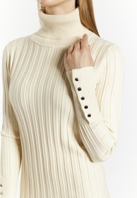 Pull en maille côtelée couleur crème à col roulé, avec des manches longues, mettant en avant des accents de boutons noirs aux poignets et un design texturé et ajusté.
