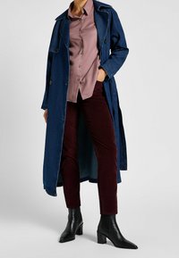 Long denim coat in dark blue with a light pink button-up shirt underneath. Burgundy corduroy pants and black heeled ankle boots complete the outfit.