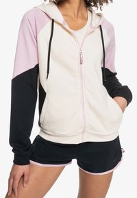 Sudadera con capucha y cremallera en colores crema, rosa y negro; presenta mangas contrastantes, bolsillos laterales y cordones en la capucha.