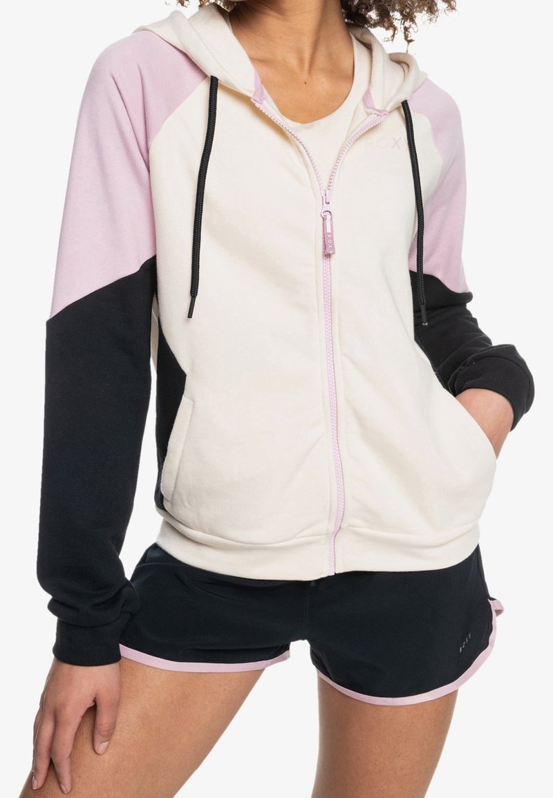 Sudadera con capucha y cremallera en colores crema, rosa y negro; presenta mangas contrastantes, bolsillos laterales y cordones en la capucha.