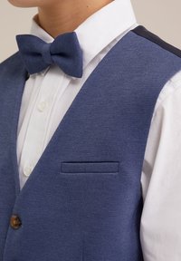 Blåtexturerad väst med en vit skjorta under, försett med en fluga och en liten ficka på vänster sida. Bruna knappdetaljer finns.