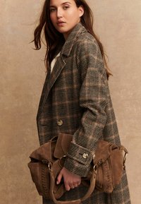 Mujer con un abrigo de cuadros marrón sosteniendo un gran bolso de ante marrón con detalles en dorado frente a un fondo neutro.